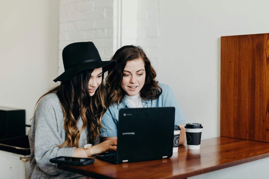 Two individuals collaborating over a laptop at a wooden table, with coffee cups nearby, in a casual workspace setting.