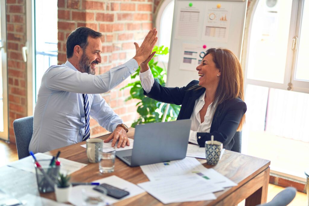 Business professionals celebrating success with high-five at a desk, surrounded by paperwork and a laptop, in a modern office setting related to tax resolution services.