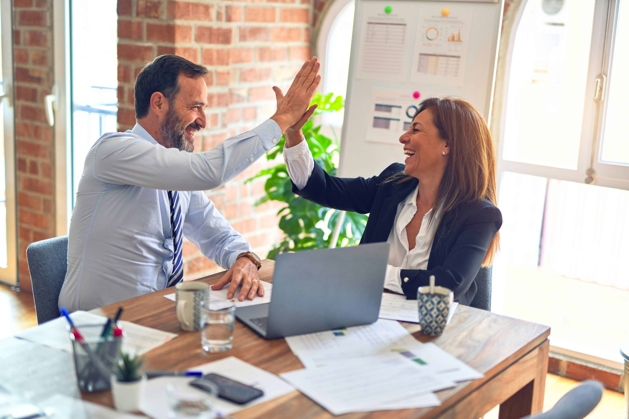 Business professionals celebrating success with high-five at a desk, surrounded by paperwork and a laptop, in a modern office setting related to tax resolution services.